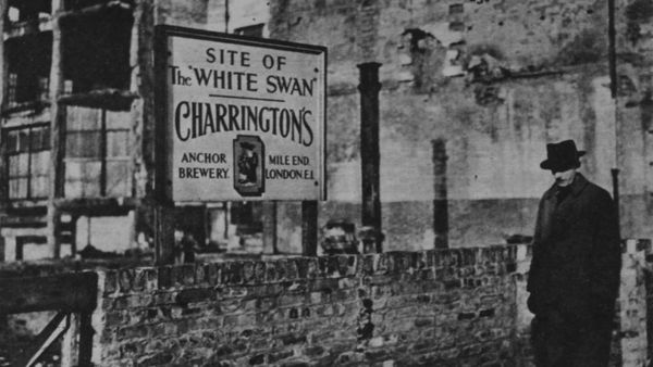 Man in a hat in front of a wall and a hand-painted sign with destroyed buildings behind.