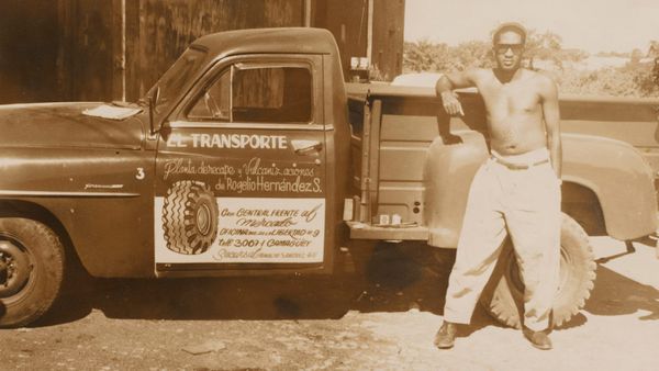 Sepia photo of a topless man resting his arm on the back of a truck that has hand-painted lettering on the door.