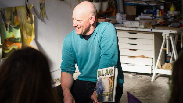 Man sitting on a stool in an artistic workshop setting, smiling and holding a magazine with 'BLAG' on the masthead.