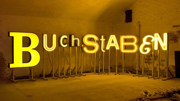 Diverse letters with yellow illumination of different forms arranged on support stands to spell 'Buchstaben'.
