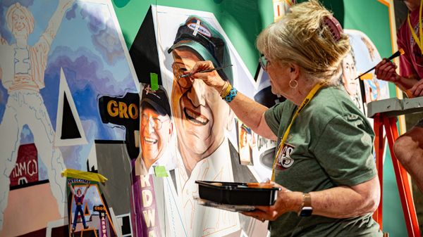View from the side of a woman painting a man's face on a mural with reference to a picture of him taped to the wall beside her.