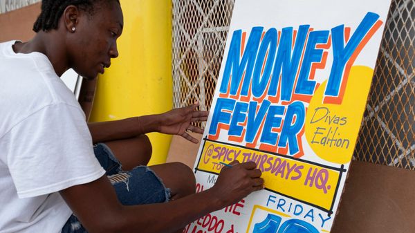 Man sat on a stool painting a sign advertising "Money Fever, Divas Edition, at Spicy Thursdays HQ."