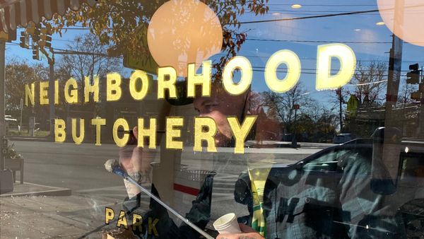 Sign painter working on the inside a window with gilded letters that read "Neighbourhood Butchery".