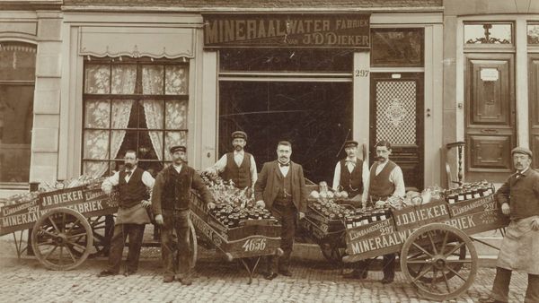 Black and white photo of men posing with loaded carts advertising "J.D. Dieker, Mineraalwater Fabriek".