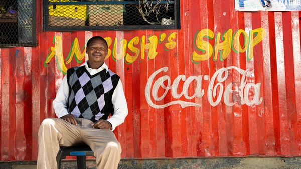 Man sat in front of a corrugated iron structure painted red and lettered with the Coca-Cola logo in white and "Ambush's Shop" in yellow above.