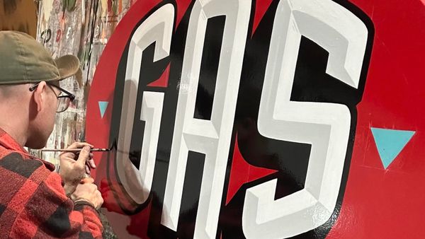 View from over the shoulder of a sign painter adding a black block shade onto large prismatic letters in white and light grey that say "Gas" set on a red background.