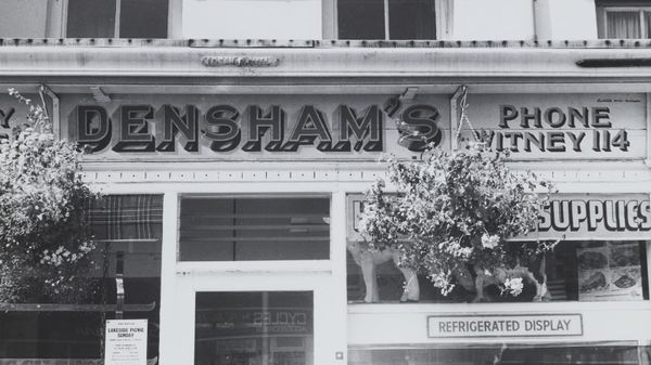 Black and white photo of a shopfront with hanging baskets and hand-painted dimensional lettering that says "Densham's" and a smaller "Phone Witney 114". 