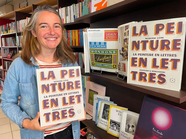 Woman holding a large book with the title in bold signwritten lettering on the cover. Another copy is displayed on the shelf of the bookshop location.