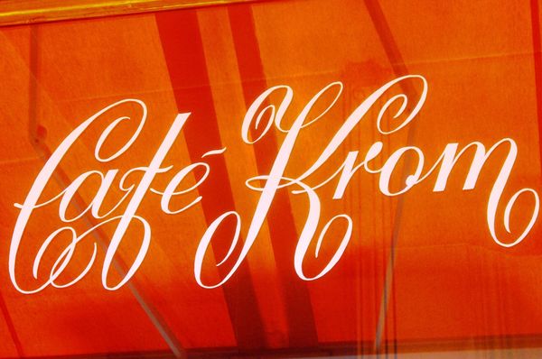 Sign painted in white by hand on a glass window reflecting an orange awning.