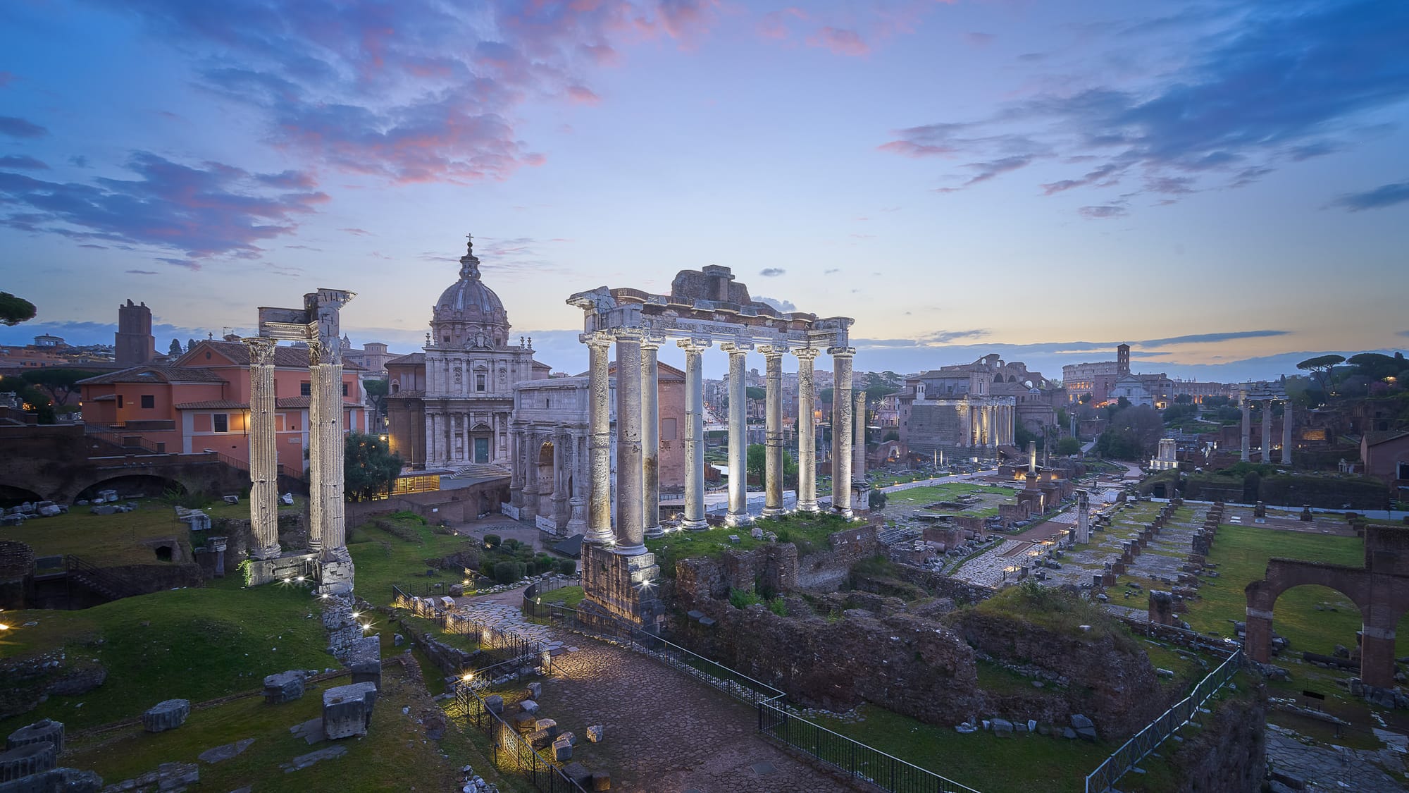 Ancient Roman Forum at sunrise with illuminated marble columns of the Temple of Saturn against pink and blue pastel sky, viewed from an elevated position with no tourists present