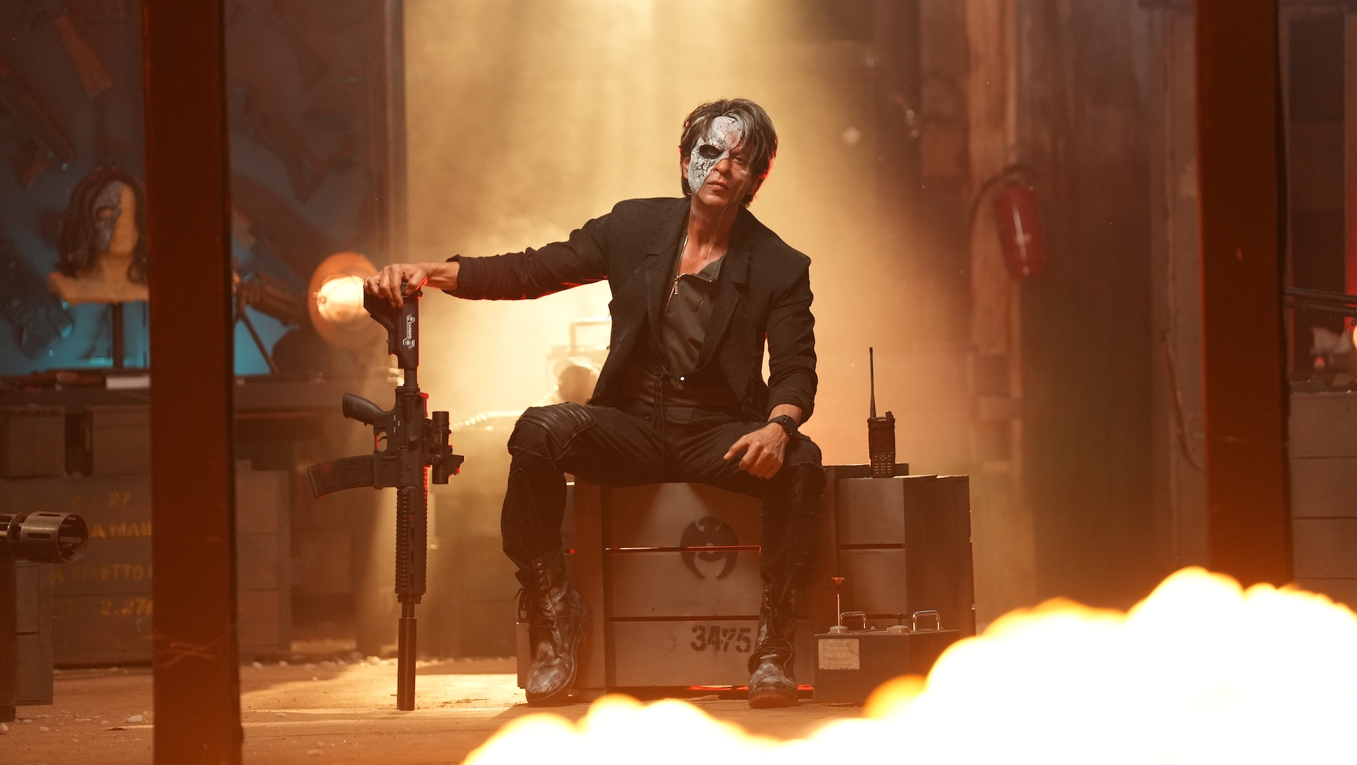 As explosions surround him, Shah Rukh Khan sits on a box and holds onto a rifle in Jawan