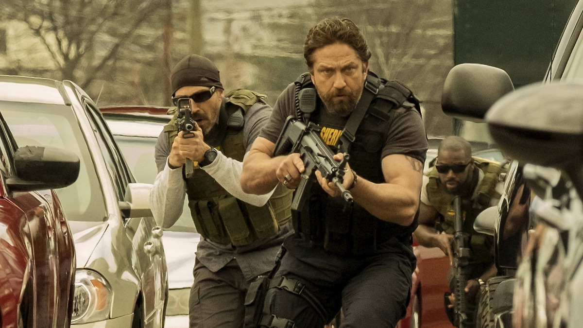 Gerard Butler charges forward with an assault rifle and a bulletproof vest, surrounded by cars, in Den of Thieves