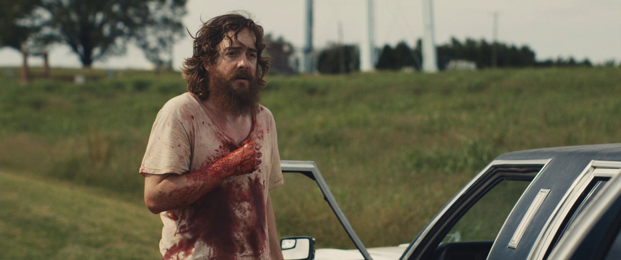 Macon Blair, standing next to a car, is covered in blood in Blue Ruin