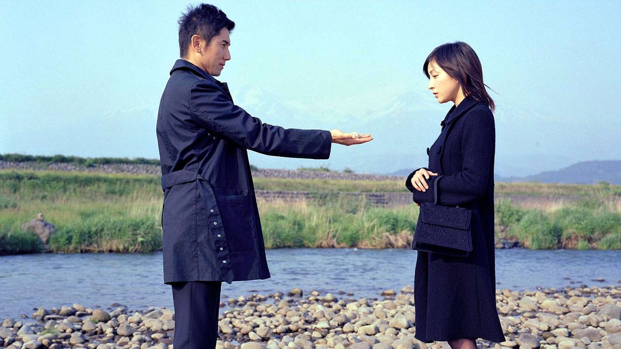 In Departures, a man holds out his outstretched palm to a woman as they stand on rocks by a river