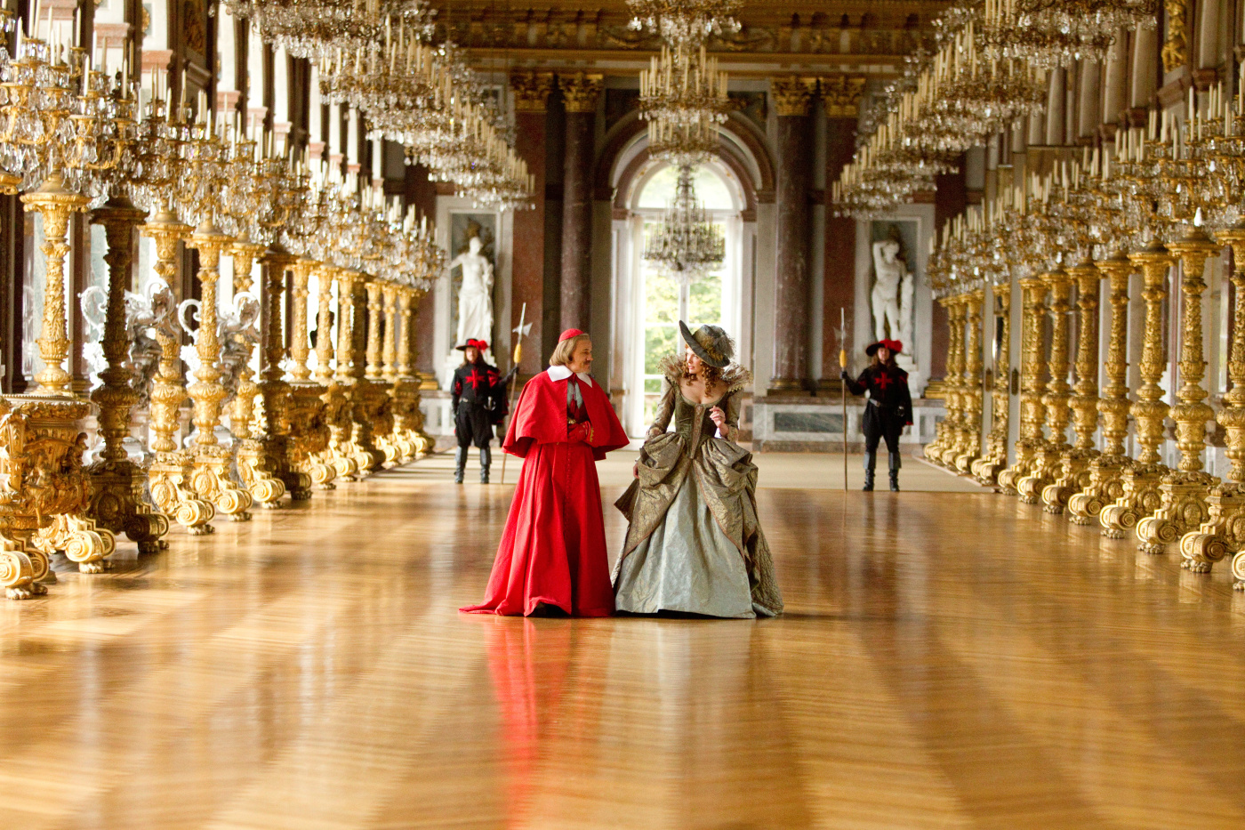 Christoph Waltz and Milla Jovovich walk down an ornate hallway with giant candelabras in The Three Musketeers