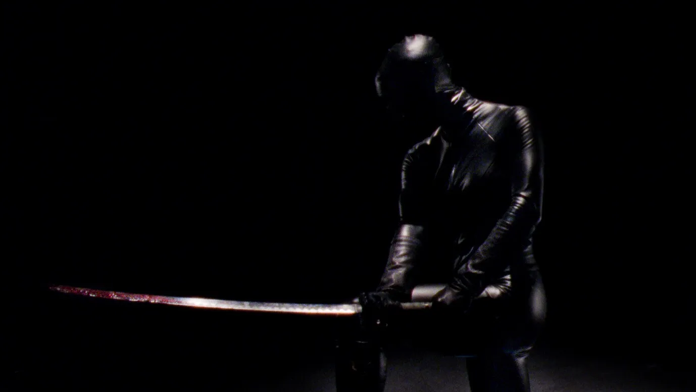 In Reflection in a Dead Diamond, a woman wearing an all-black leather skintight outfit covering her head and face swings a bloody sword