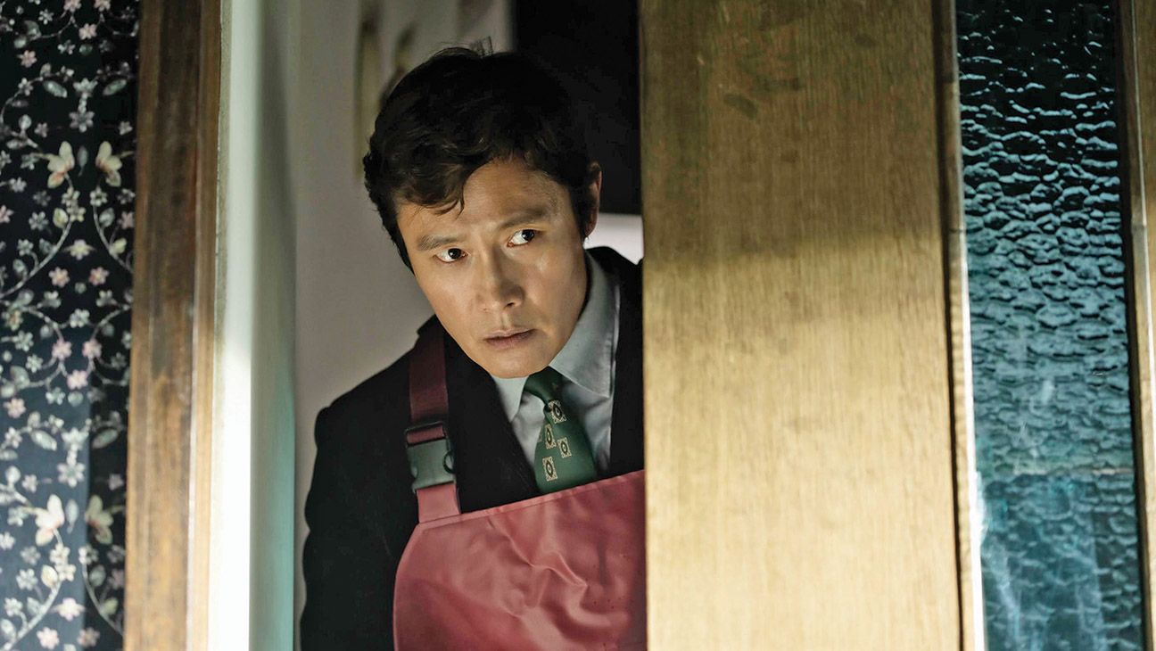 Lee Byung-hun, wearing an apron, peers through an open doorway in No Other Choice