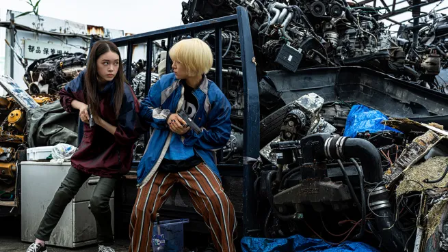 In Baby Assassins 2, Maharo and Chisato hold pistols and get ready to fight in a junkyard
