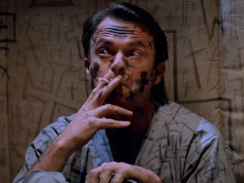 Sam Neill, crosses all over his face and clothes, smokes a cigarette in In the Mouth of Madness