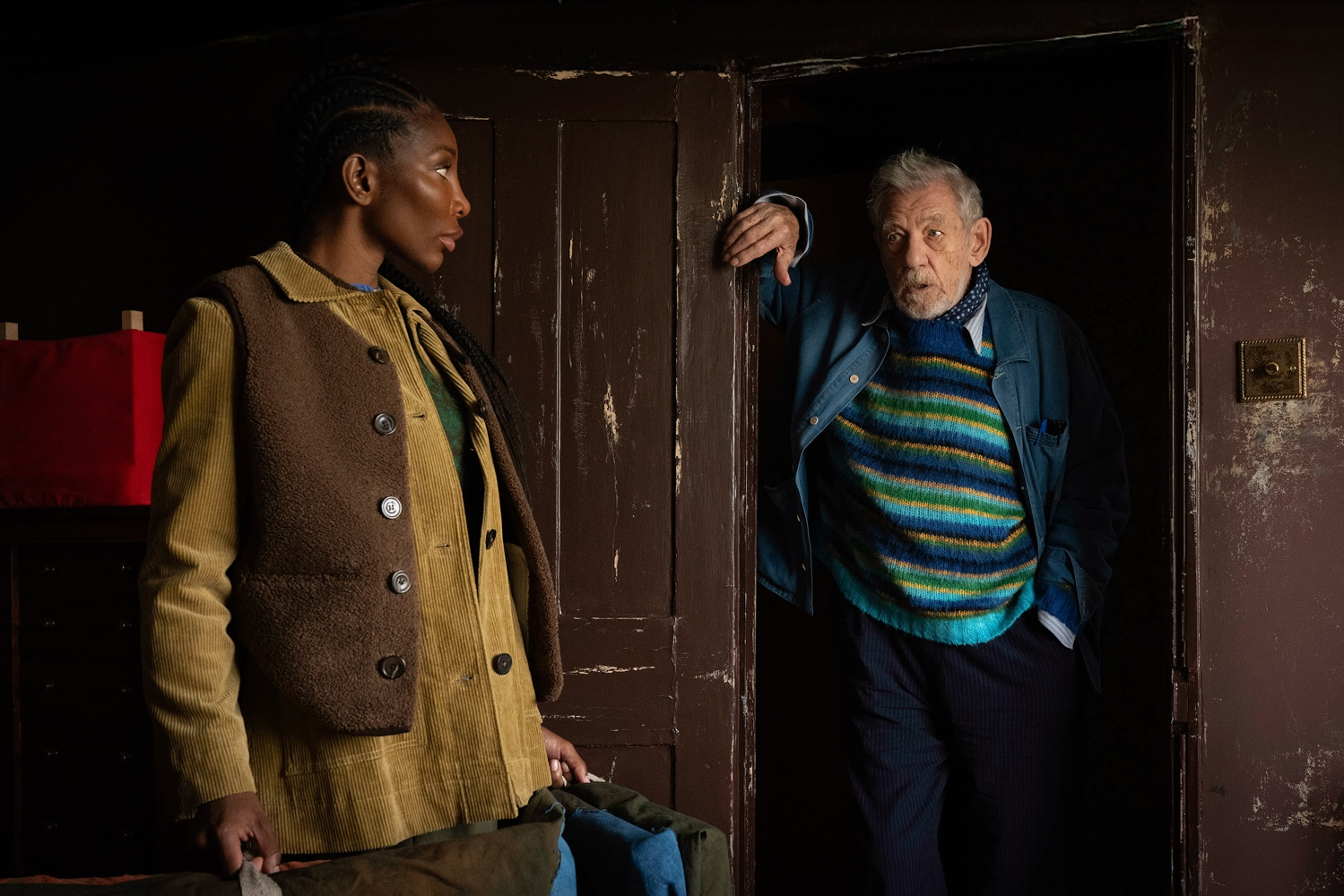 Ian McKellen leaning in a doorway as Michaela Coel carries bags in The Christophers