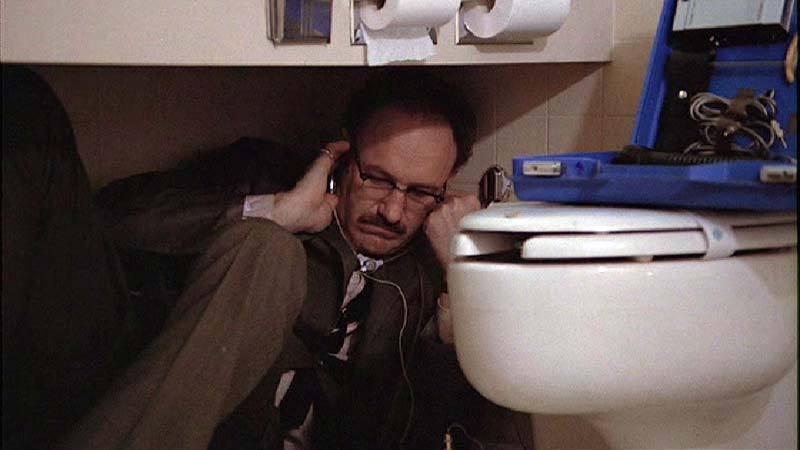 Gene Hackman folded under a bathroom sink, next to a toilet, listening intently with surveillance equipment in The Conversation