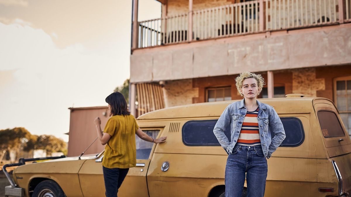 Julia Garner stands in front of an old car outside of an old hotel in The Royal Hotel