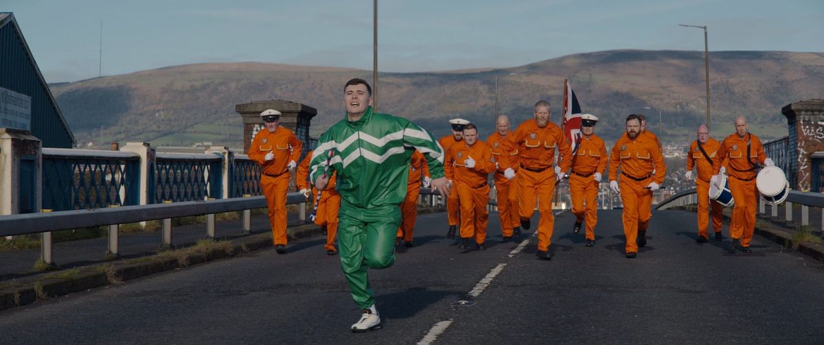 Kneecap's Mo Chara, wearing a green tracksuit, runs away from a bunch of Irish Loyalists wearing orange in Kneecap