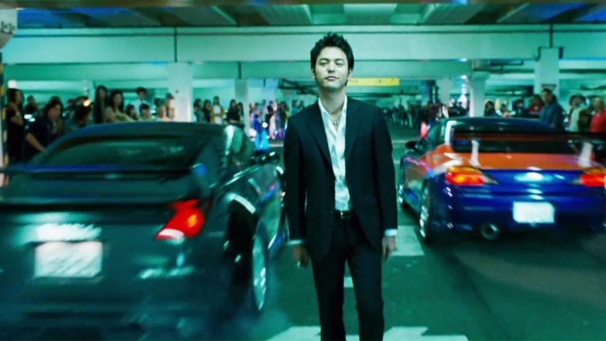 Drift cars race by a man in a parking lot as spectators watch in Tokyo Drift