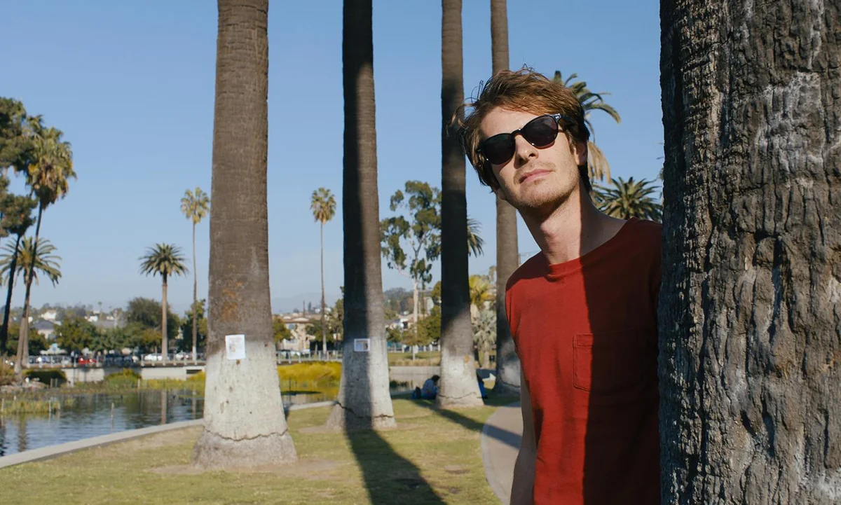 Andrew Garfield, looking like he hasn't slept in weeks, peering out from behind a palm tree while wearing sunglasses in Under the Silver Lake
