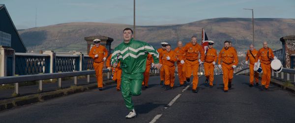 Kneecap's Mo Chara, wearing a green tracksuit, runs away from a bunch of Irish Loyalists wearing orange in Kneecap