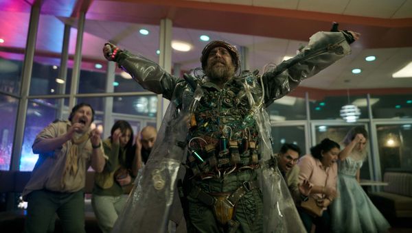 Sam Rockwell wearing what appears to be a bomb vest and a clear rain jacket while people cower behind him in a diner in Good Luck, Have Fun, Don't Die