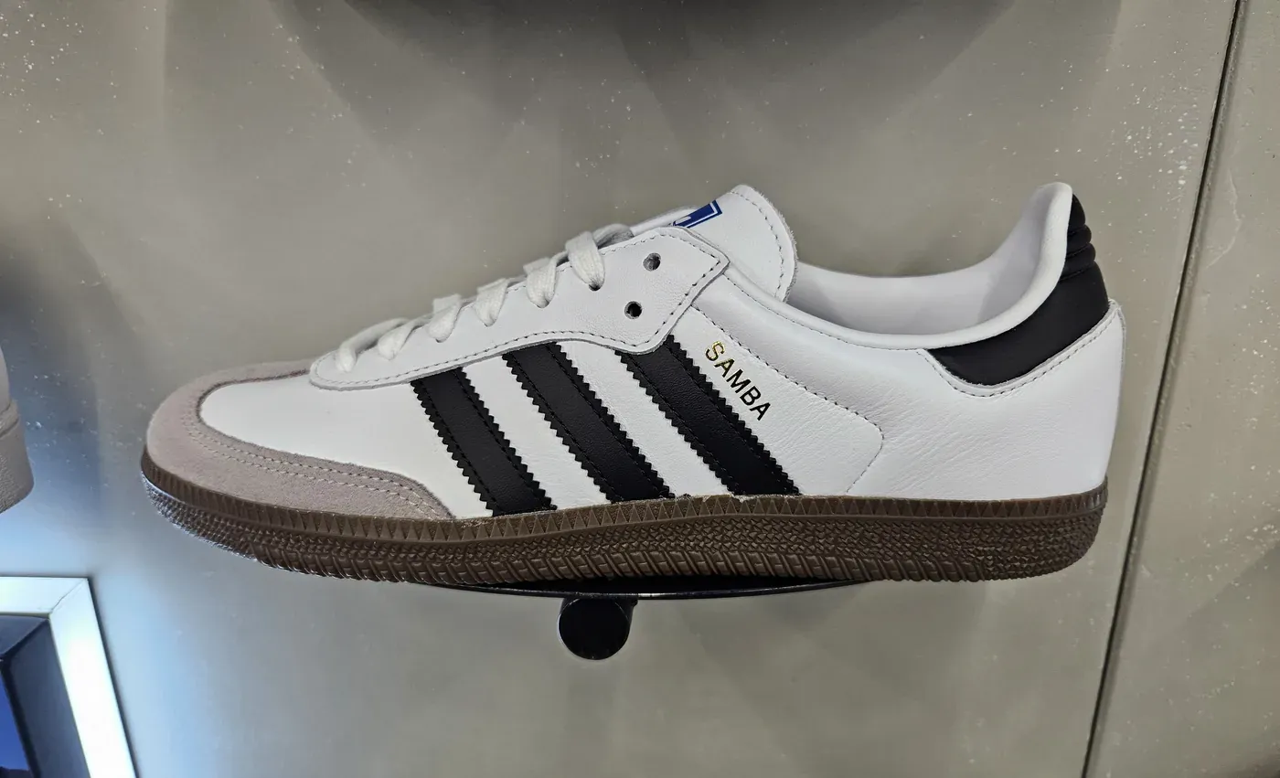 White adidas Samba OG sneaker with black stripes and a gum sole displayed on a wall shelf