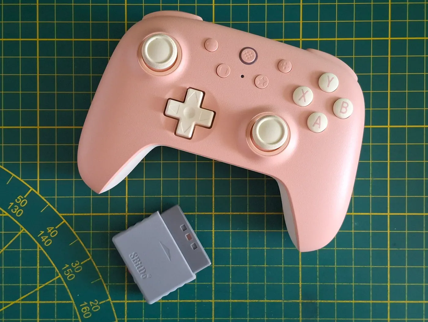 Pink 8BitDo Ultimate 2C wireless controller next to a grey USB receiver on a green cutting mat