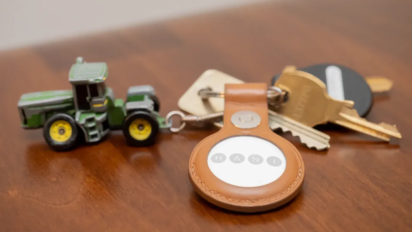 Apple AirTag in a tan leather key ring holder beside a set of keys and a toy tractor on a wooden table