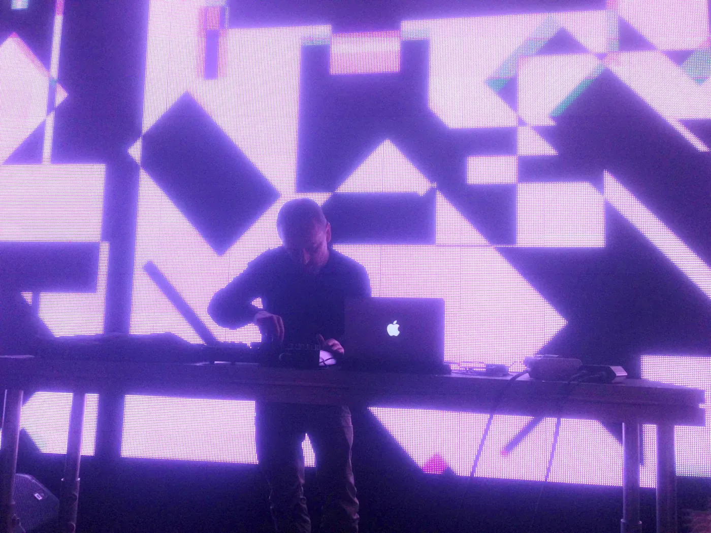 Mark Bell of LFO performing behind a table of gear in front of a purple geometric light projection
