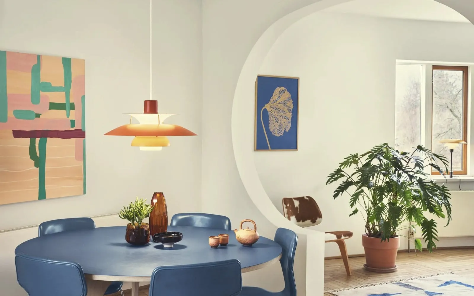 A Louis Poulsen PH 5 pendant lamp in orange tones hanging over a blue dining table in a bright room