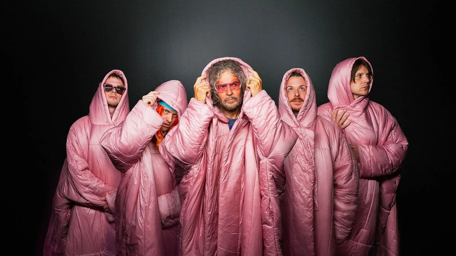The Flaming Lips in hooded pink padded coats against a black backdrop, Wayne Coyne in the center wearing pink glasses