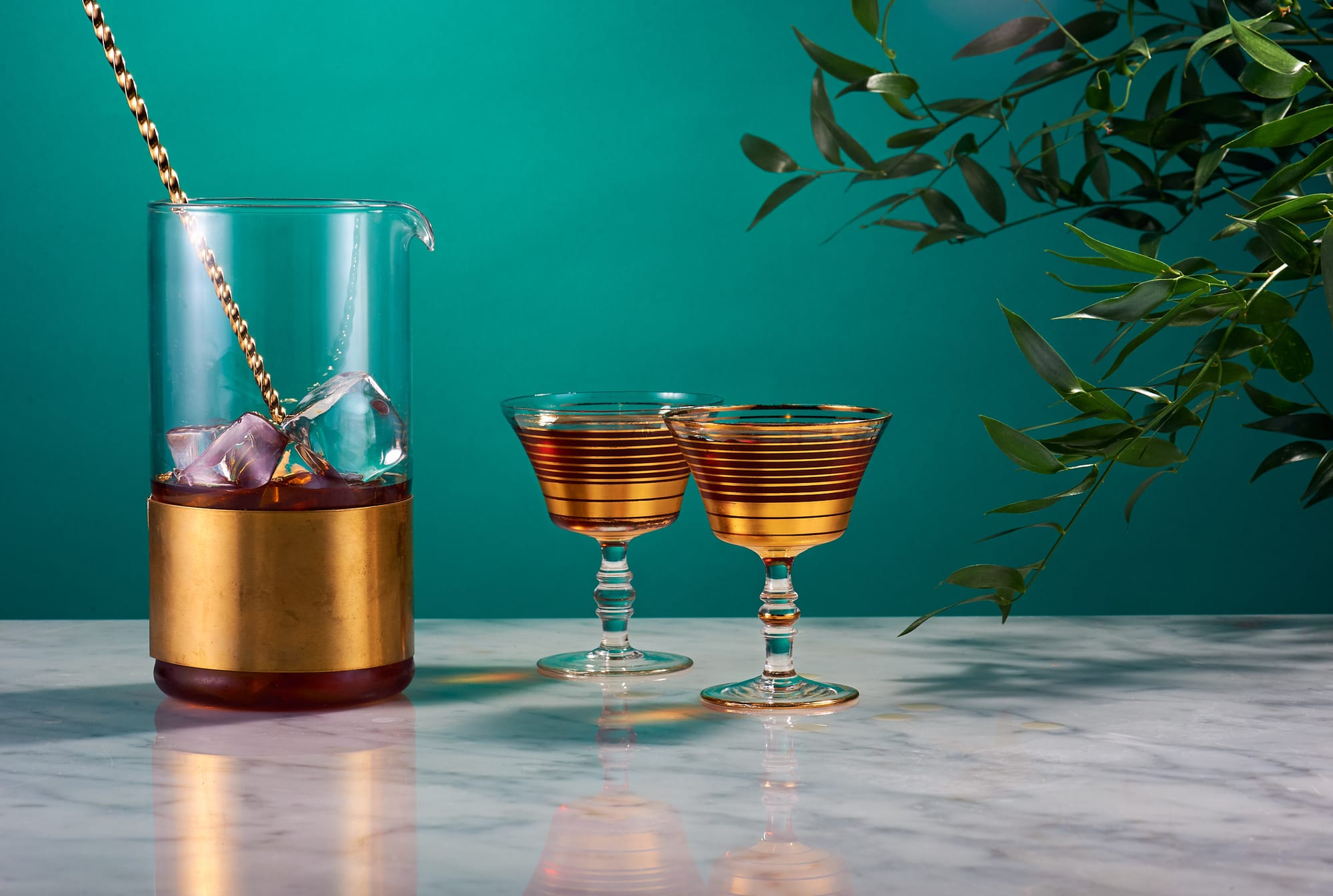 Elegant cocktail setup with gold-banded mixing glass and striped coupe glasses filled with amber liquid, against a teal background with olive branches