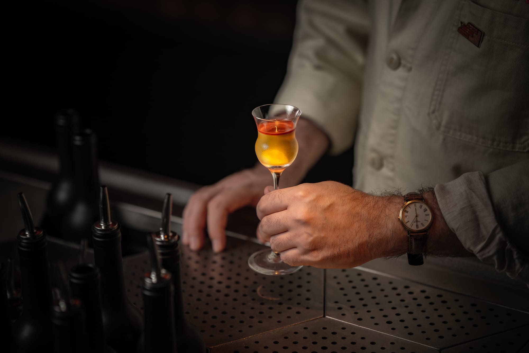 Bartender in white shirt holding a layered amber cocktail in a small stemmed glass, with bar tools visible in the background
