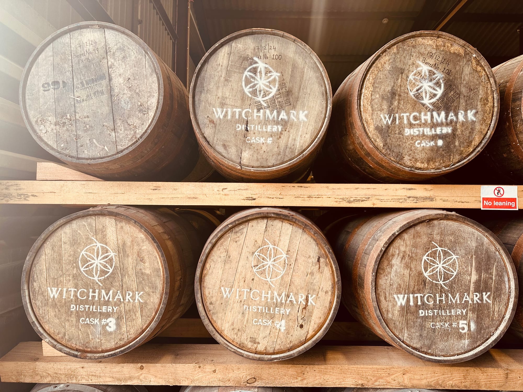 Photograph of six wooden casks arranged in two rows of three, seen from end on. The barrel heads bear the Witchmark distillery name and logo and on the bottom row are numbered: 3, 4 and 5. (The one top right is cask number 1 but that info is on the end facing away from us.) There is a small red and white warning sign on the wooden cask rack forbidding us from leaning on it. As if we would!