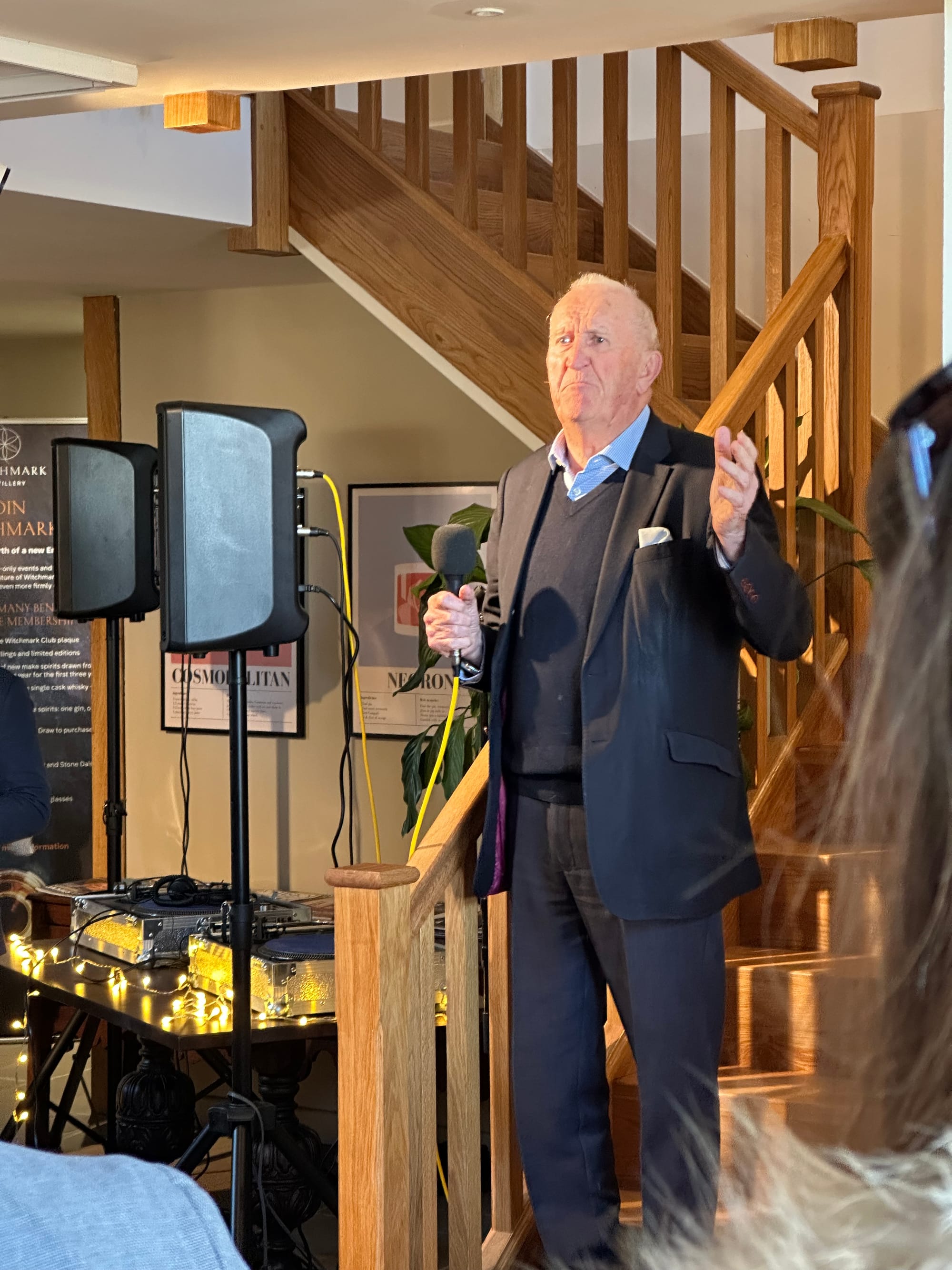 A not especially flattering photograph of a man around 80 years old, dressed in a navy blue suit (with magenta silk lining), a navy v neck jumper and blue shirt. He is standing on some wooden stairs, the first or second step. He holds a microphone in his right hand but doesn't appear to be using it, rather he holds it near mid-chest level. He is gesticulating with his left hand, which is open. His facial expression looks grumpy but that is most likely just unfortunate timing, as he was mid speech and faces do weird things when we talk. There is a table behind him with DJ deck, and two speakers on stands.