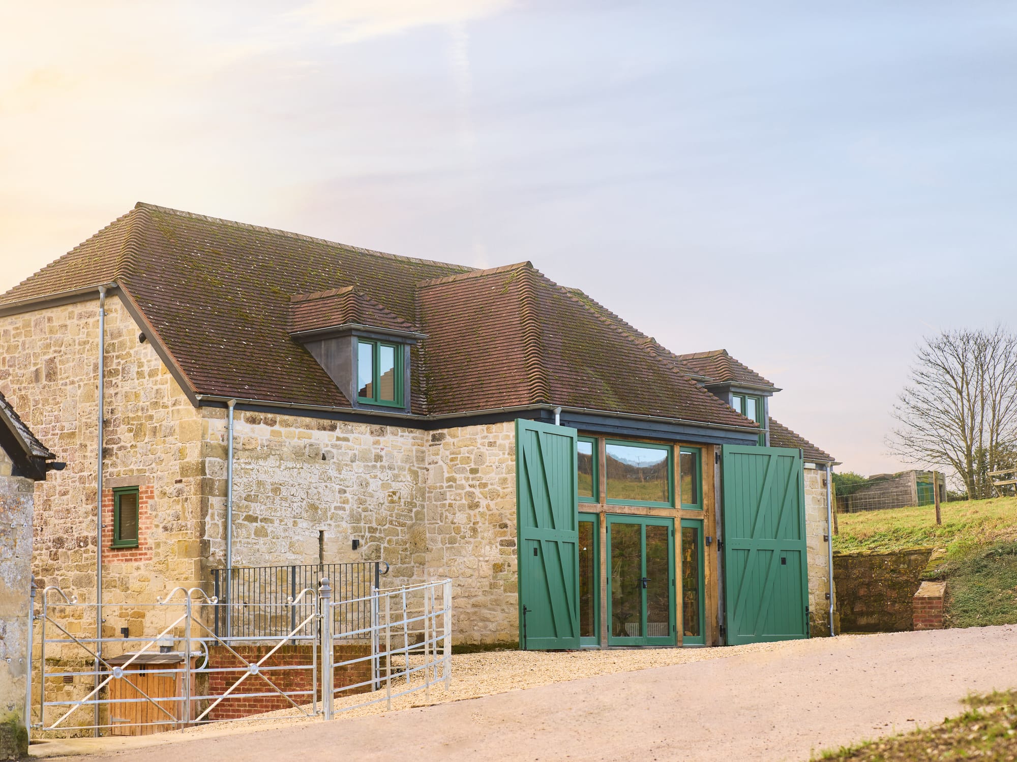A barn dating from the 1600s made of stone, with large wooden doors painted sage green, and a roof of clay tiles in which there are two dormer windows, one either side of the main doors. Within the opening of the main doors there are windows and two smaller glass doors.