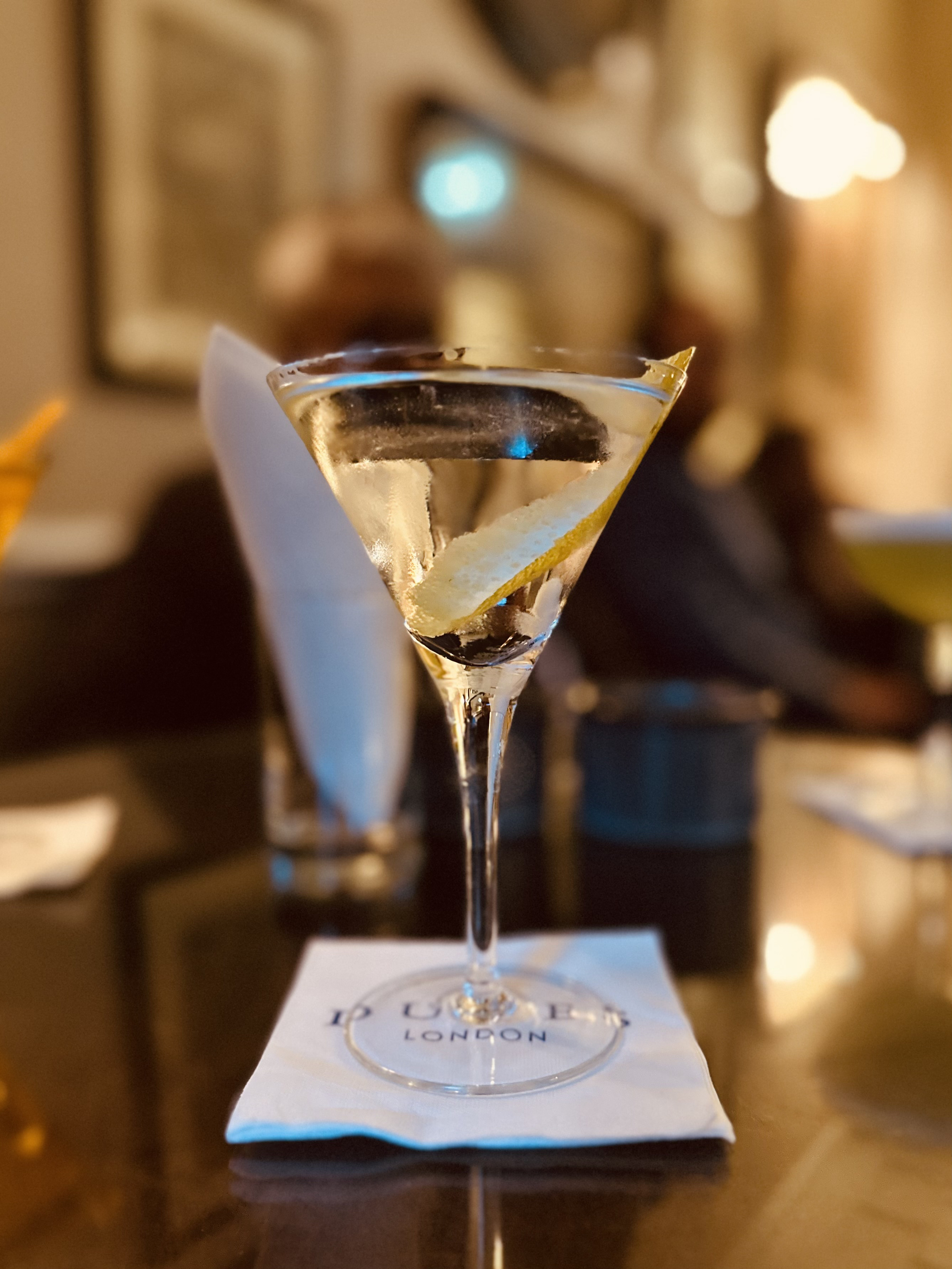 A photograph of a martini cocktail in the classic Y-shaped glass, garnished with a twist of Amalfi lemon. It stands atop a paper cocktail napkin from which we can tell this particular martini was served at Dukes bar in London.