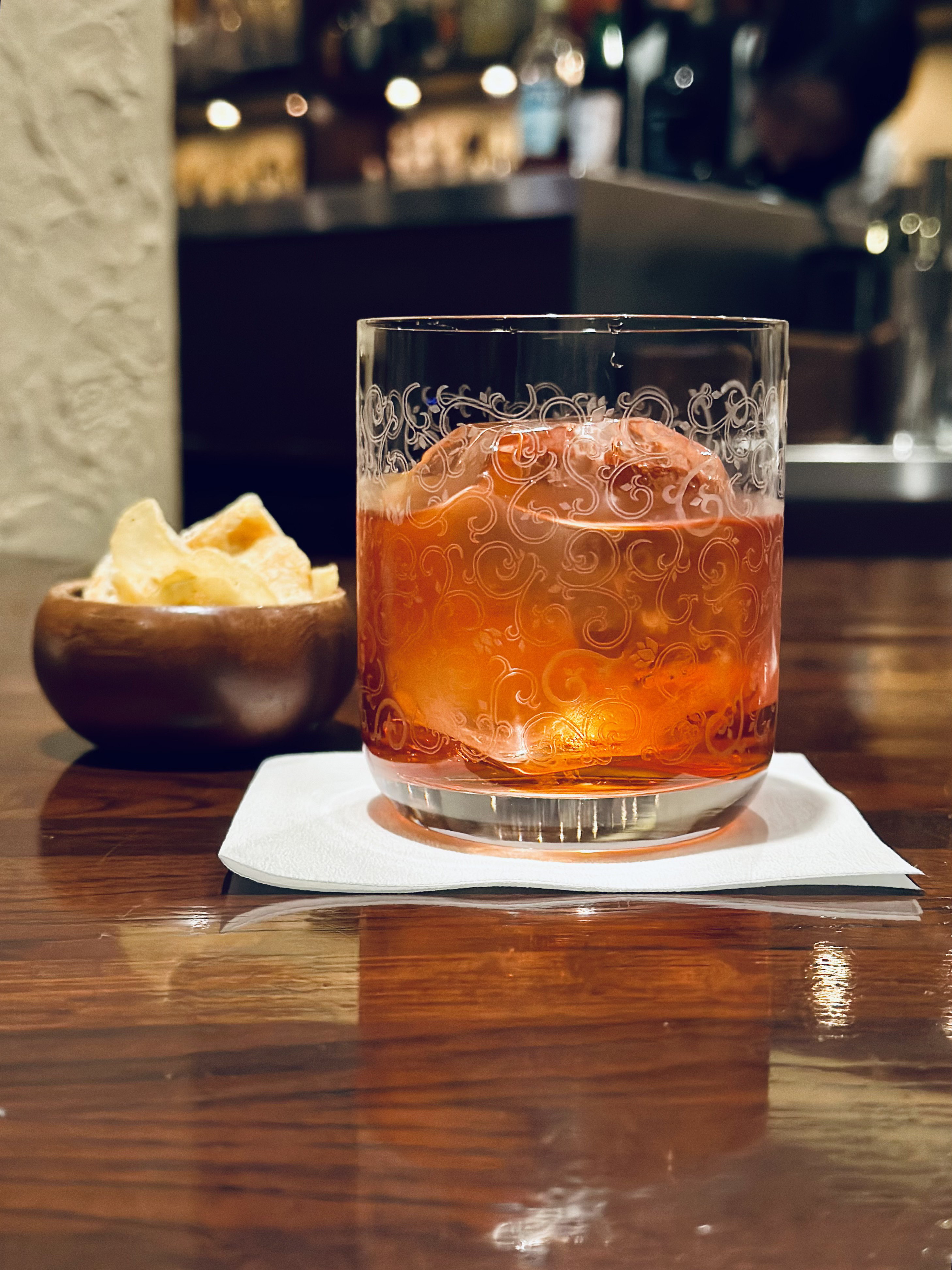 Photograph of a cocktail on a bar napkin next to a small bowl of snacks. It is in a rocks glass with swirls of decorative etching. It is reddish-orange and there is a large rock of ice that takes up much of the glass. In the background is an out-of-focus view of a city street at night.