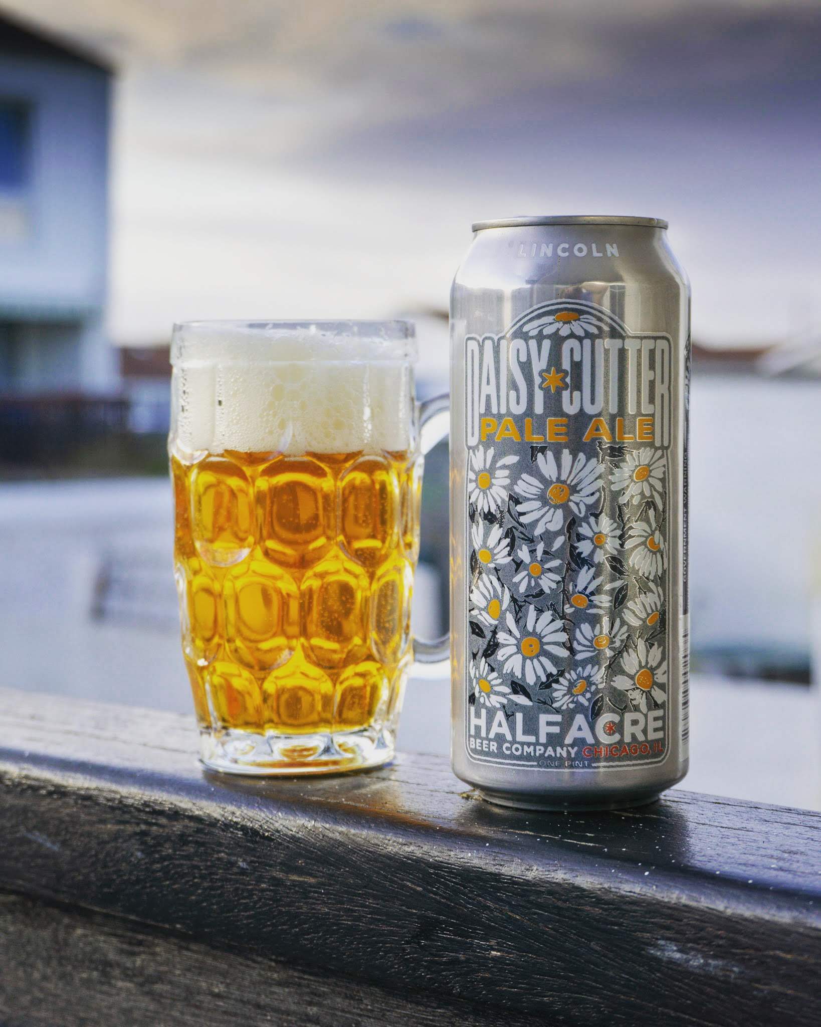 A small dimpled glass of golden pale ale with a foamy head sits beside a silver can labelled “Daisy Cutter Pale Ale, Half Acre Beer Company, Chicago, IL,” against a soft, cloudy outdoor backdrop.