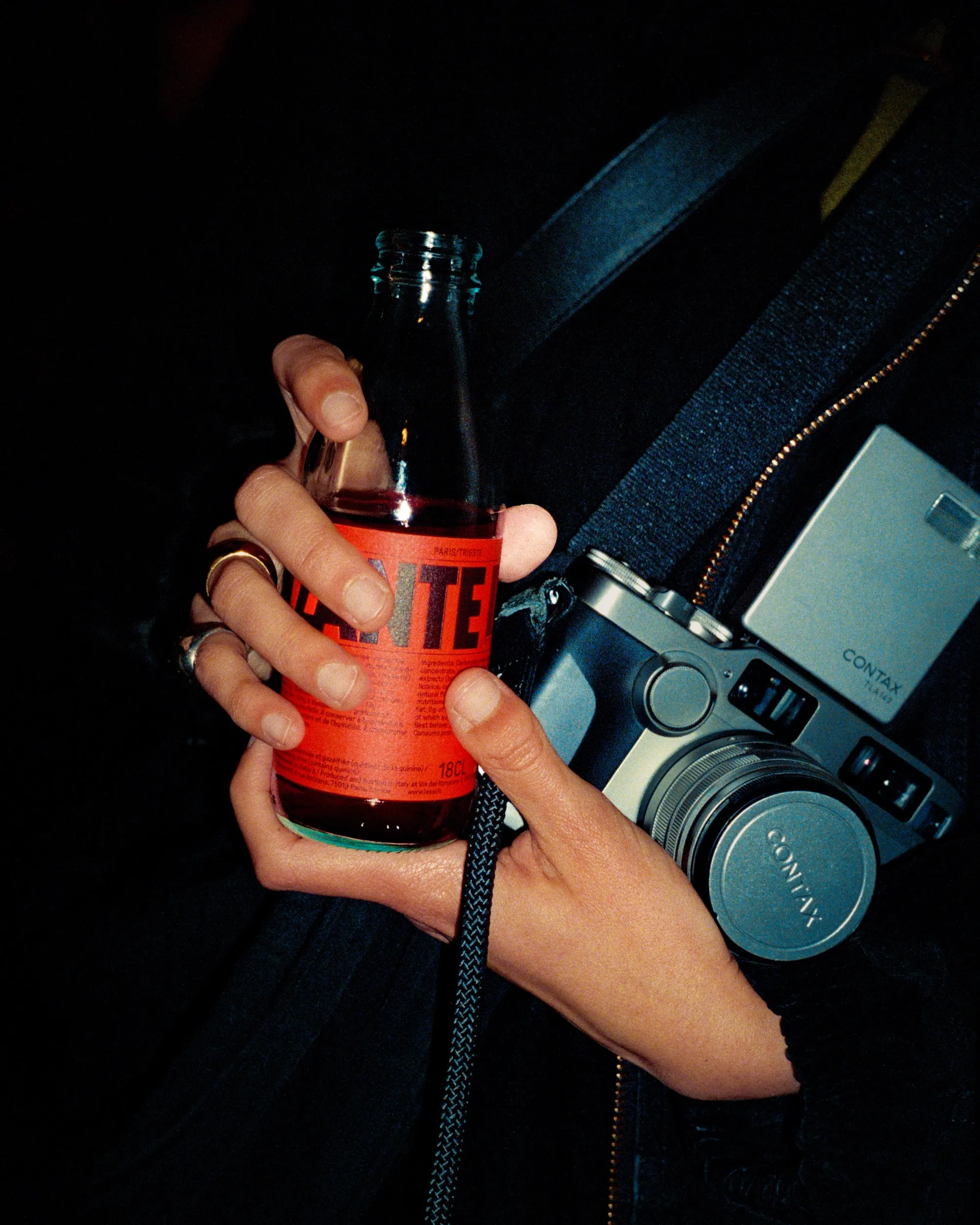Close up of a pair of hands - young, maybe male? - holding a bottle of IESSI Il Frizzante and a retro Contax camera