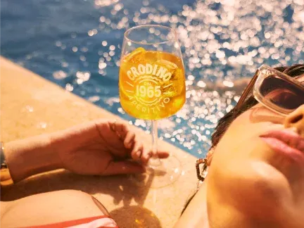 Close up of a glass of Crodino next to a woman sunbathing by a pool - we see only part of her face and shoulder, plus one hand draped onto the base of the glass