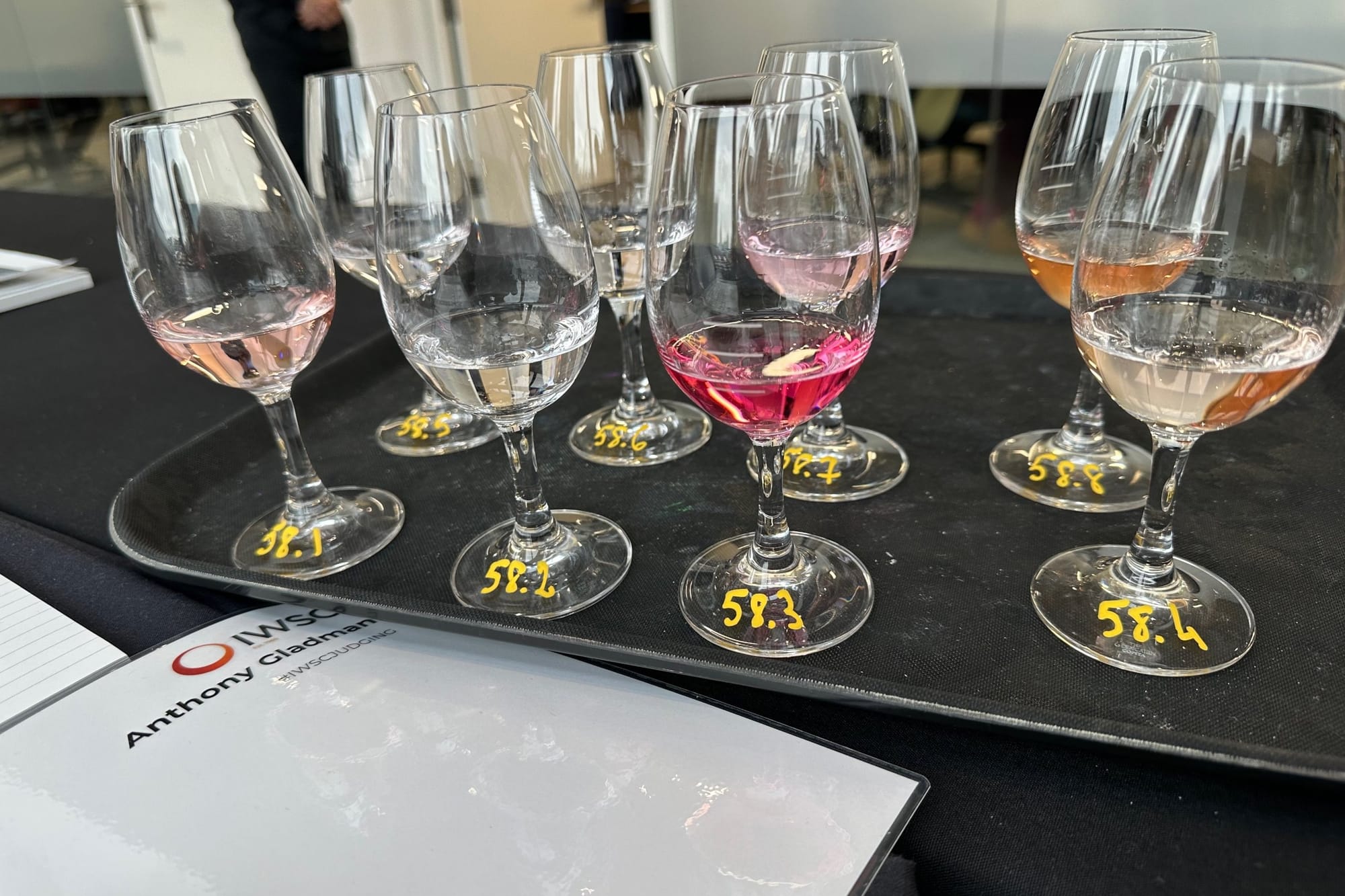 A tray of eight tasting glasses of pink and pale spirits, each foot numbered by hand from 58.1 to 58.8, beside a judge’s folder printed with the IWSC logo and the name Anthony Gladman.