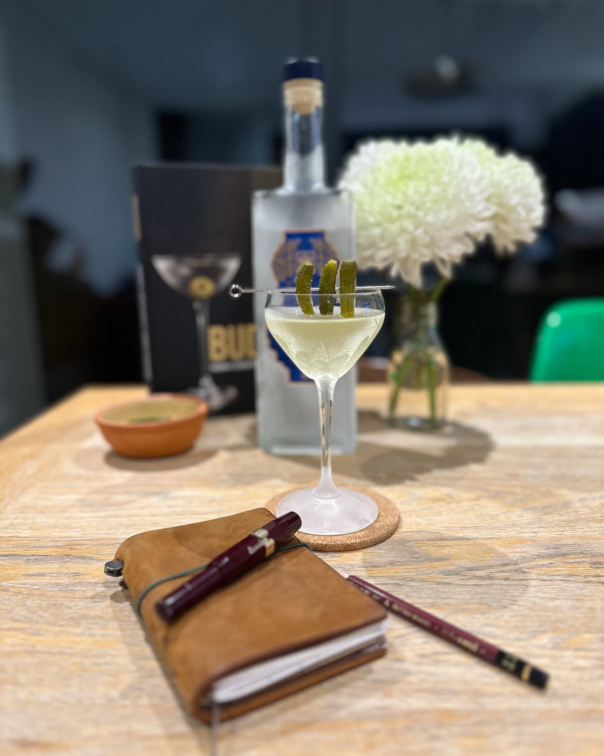 A martini in a coupe garnished with three cornichons on a pick, in front of a bottle of vodka and a vase of white chrysanthemums, with a leather notebook, fountain pen and pencil on the wooden table in the foreground.