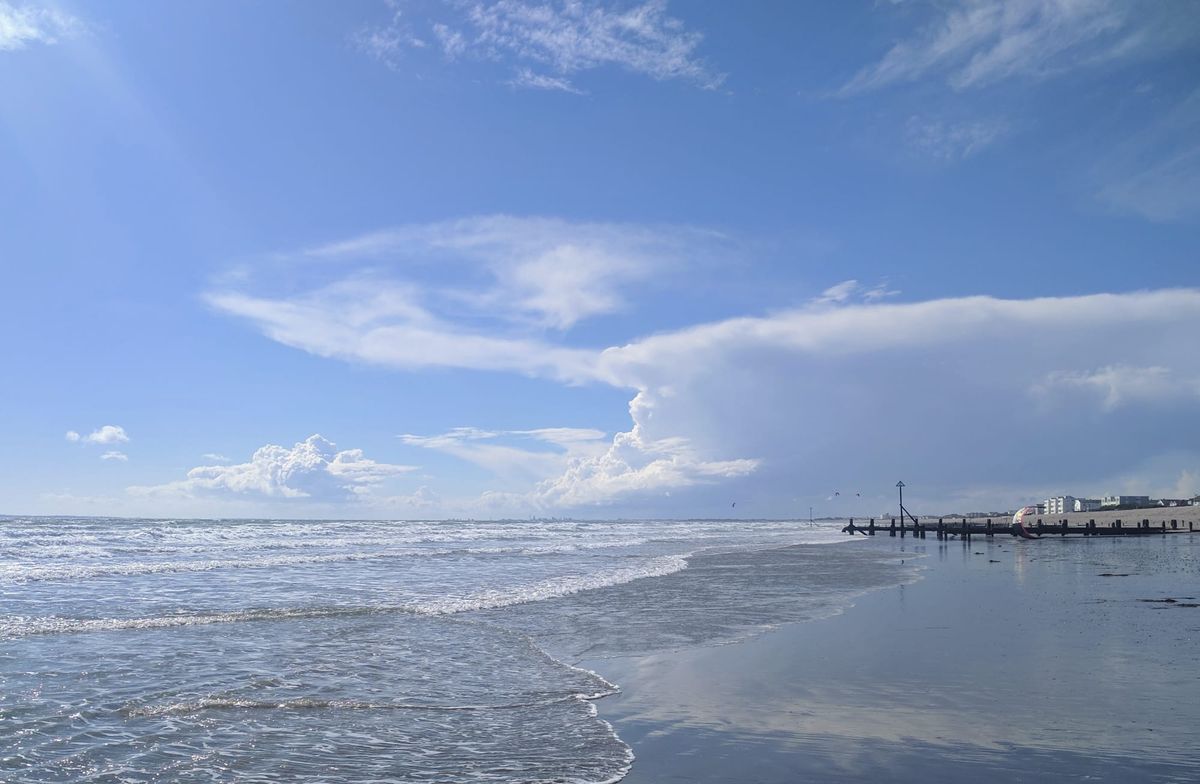 Calm waves lap against a wide sandy beach under a bright blue sky streaked with wispy and towering white clouds, with wooden groynes and small seaside buildings in the distance.